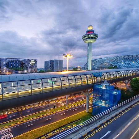 Crowne Plaza Changi Airport, An Ihg Hotel Singapore Exterior photo The photo depicts an airport scene, likely featuring a modern terminal structure with a distinctive, futuristic design. In the foreground, there is a pedestrian bridge or walkway, along with roads leading to the terminal. A control tower can be seen 