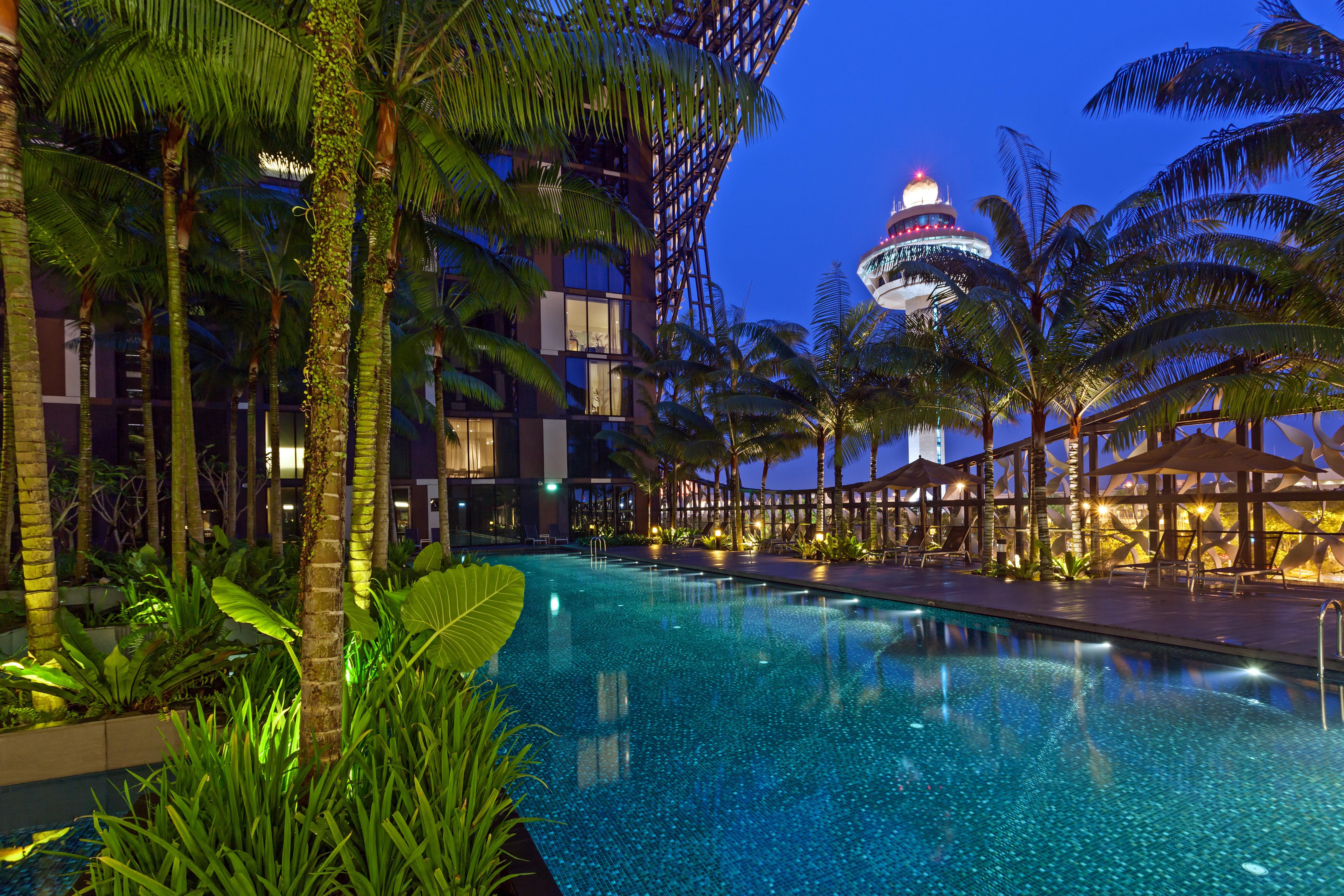 Crowne Plaza Changi Airport, An Ihg Hotel Singapore Exterior photo The photo depicts a luxurious outdoor swimming pool surrounded by lush palm trees and vibrant greenery. The scene is set during the evening, with soft lighting illuminating the water and plants. In the background, you can see a modern building struct