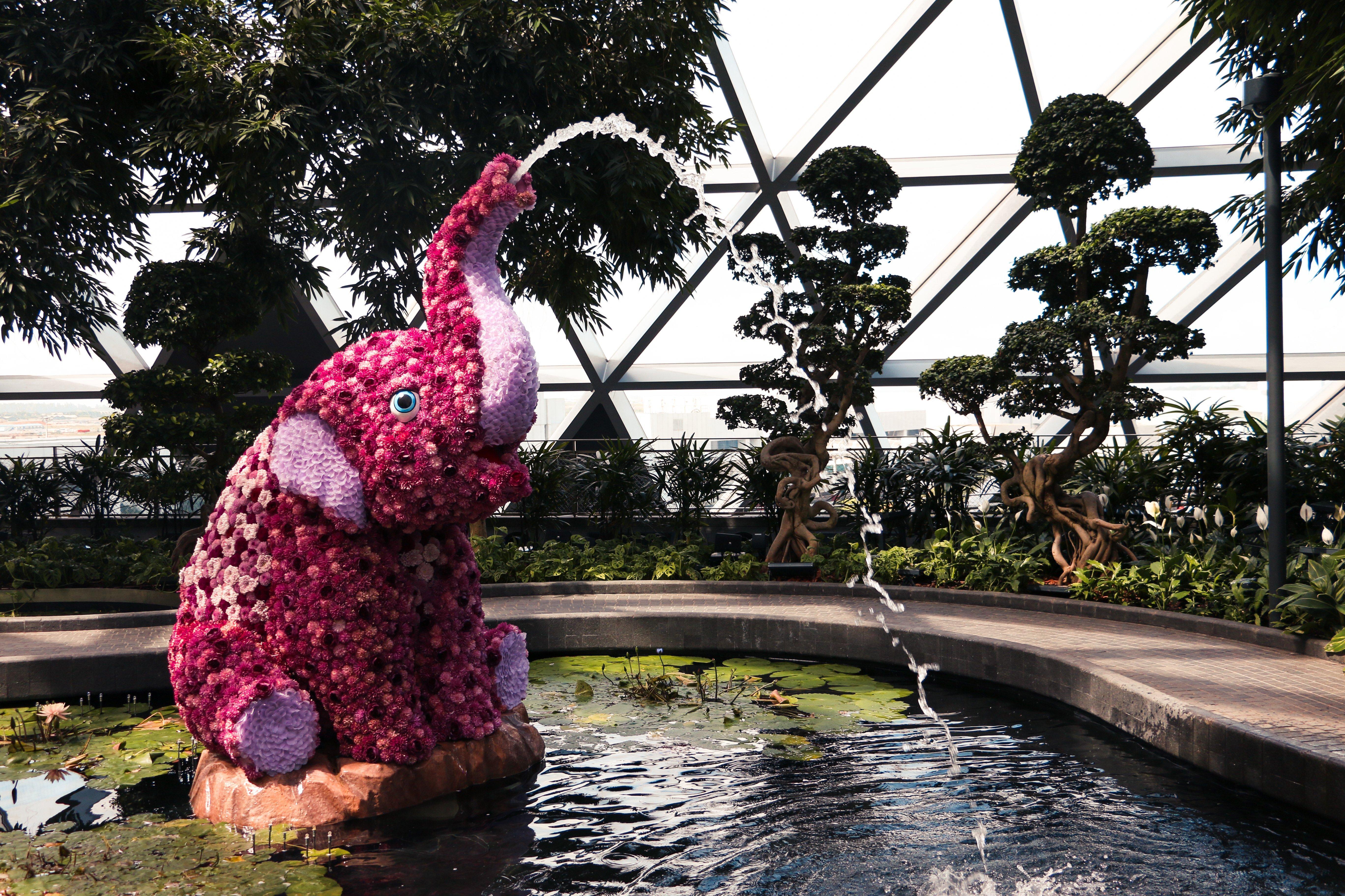 Crowne Plaza Changi Airport, An Ihg Hotel Singapore Exterior photo The photo depicts a decorative elephant sculpture made of flowers, positioned near a pond. The elephant is sitting on its hind legs, with its trunk raised high, creating a water fountain effect. Surrounding the elephant are lush green plants and tree