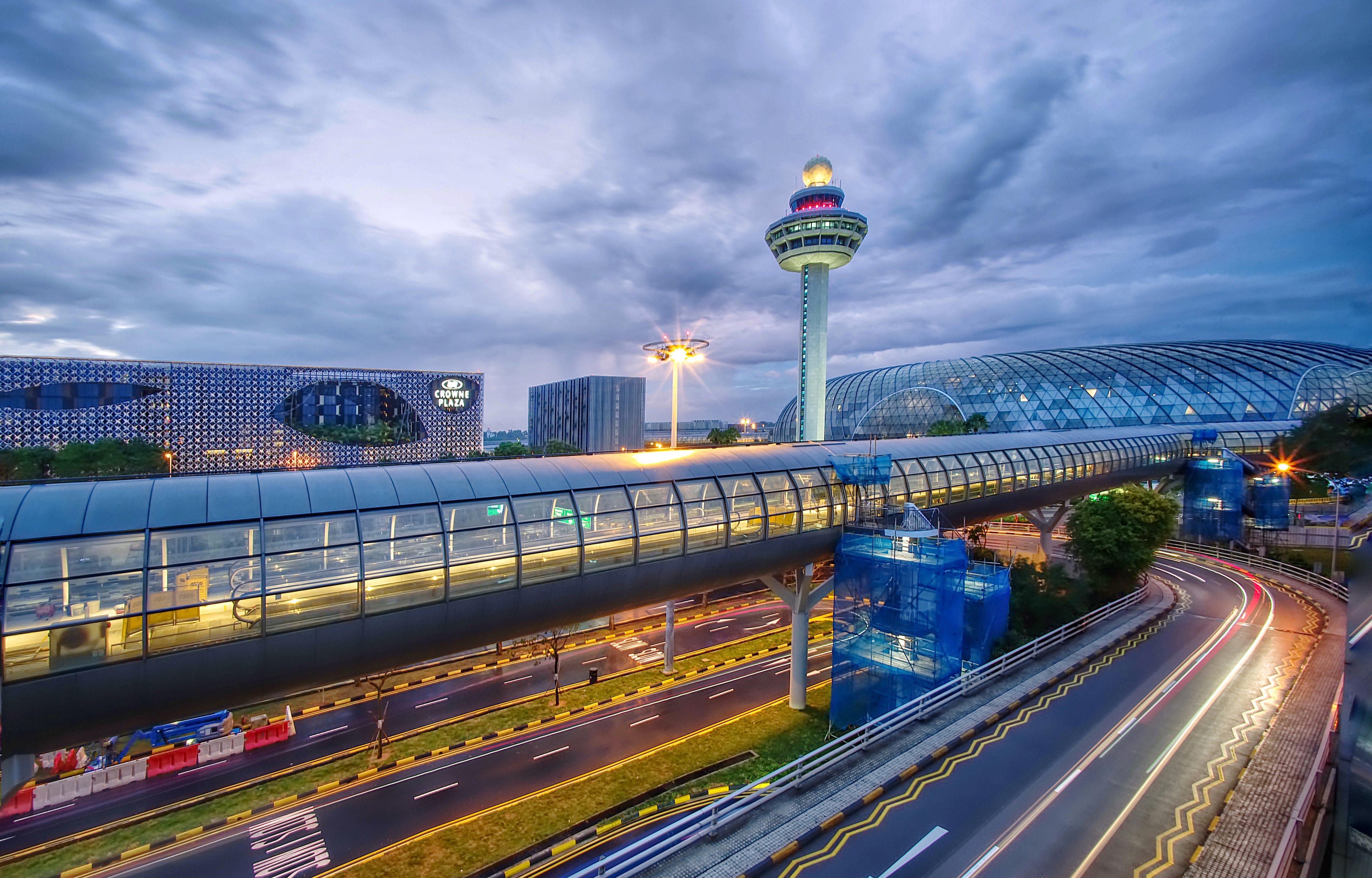 Crowne Plaza Changi Airport, An Ihg Hotel Singapore Exterior photo The photo depicts an airport scene, likely featuring a modern terminal structure with a distinctive, futuristic design. In the foreground, there is a pedestrian bridge or walkway, along with roads leading to the terminal. A control tower can be seen 