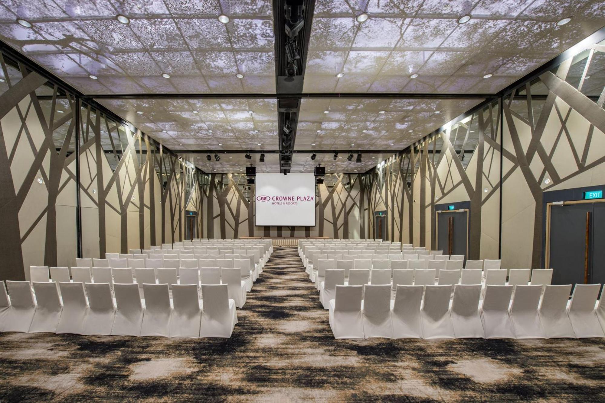 Crowne Plaza Changi Airport, An Ihg Hotel Singapore Exterior photo The photo shows a modern conference or event space. The room features rows of white chairs arranged in a theater-style layout, facing a large screen with a simple logo. The ceiling is designed to resemble a natural setting, with images that suggest t