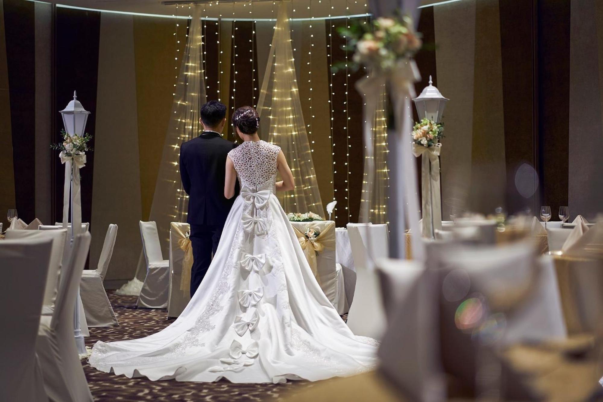 Crowne Plaza Changi Airport, An Ihg Hotel Singapore Exterior photo The photo features a couple at a wedding venue. The bride, wearing an elegant white gown with intricate details and a long train, is seen from behind. The groom stands beside her, dressed in a dark suit. The background showcases a beautifully decorat