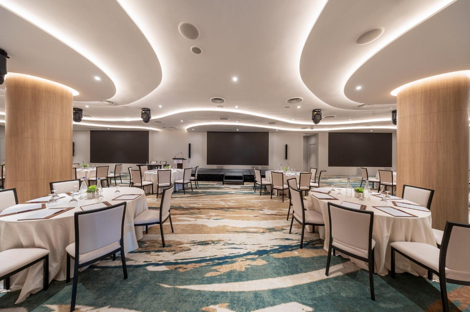 Crowne Plaza Changi Airport, An Ihg Hotel Singapore Exterior photo The photo shows a modern banquet or conference room. The room features elegantly arranged round tables with white tablecloths and chairs. The flooring has an artistic design that combines various shades of blue and beige. The ceiling is accentuated w