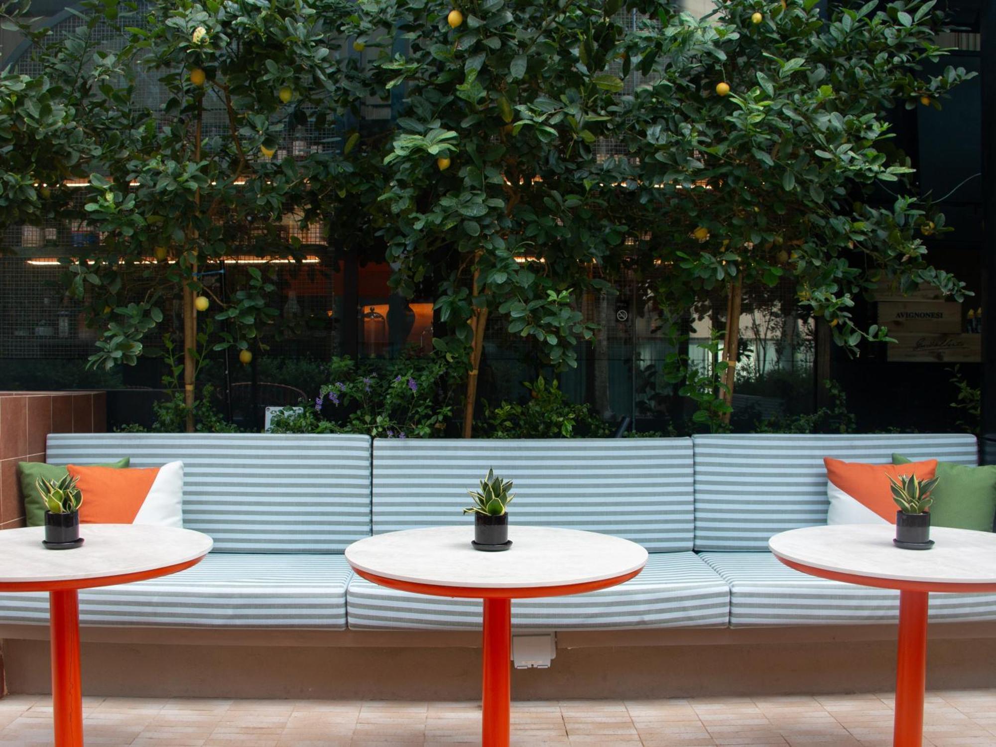 Crowne Plaza Changi Airport, An Ihg Hotel Singapore Exterior photo The photo shows a cozy seating area featuring a long, light blue bench with colorful cushions in orange and green. In front of the bench, there are three round tables with bright red bases and small plants in black pots on each table. The background 