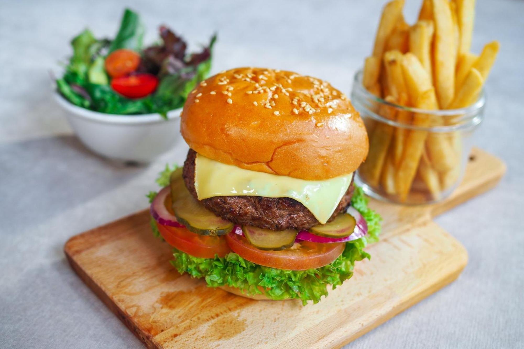 Crowne Plaza Changi Airport, An Ihg Hotel Singapore Exterior photo The photo shows a delicious looking hamburger with a sesame seed bun. The burger is layered with a beef patty, melted cheese, lettuce, tomato slices, pickles, and red onion. It is presented on a wooden cutting board. Next to the burger, there is a sm