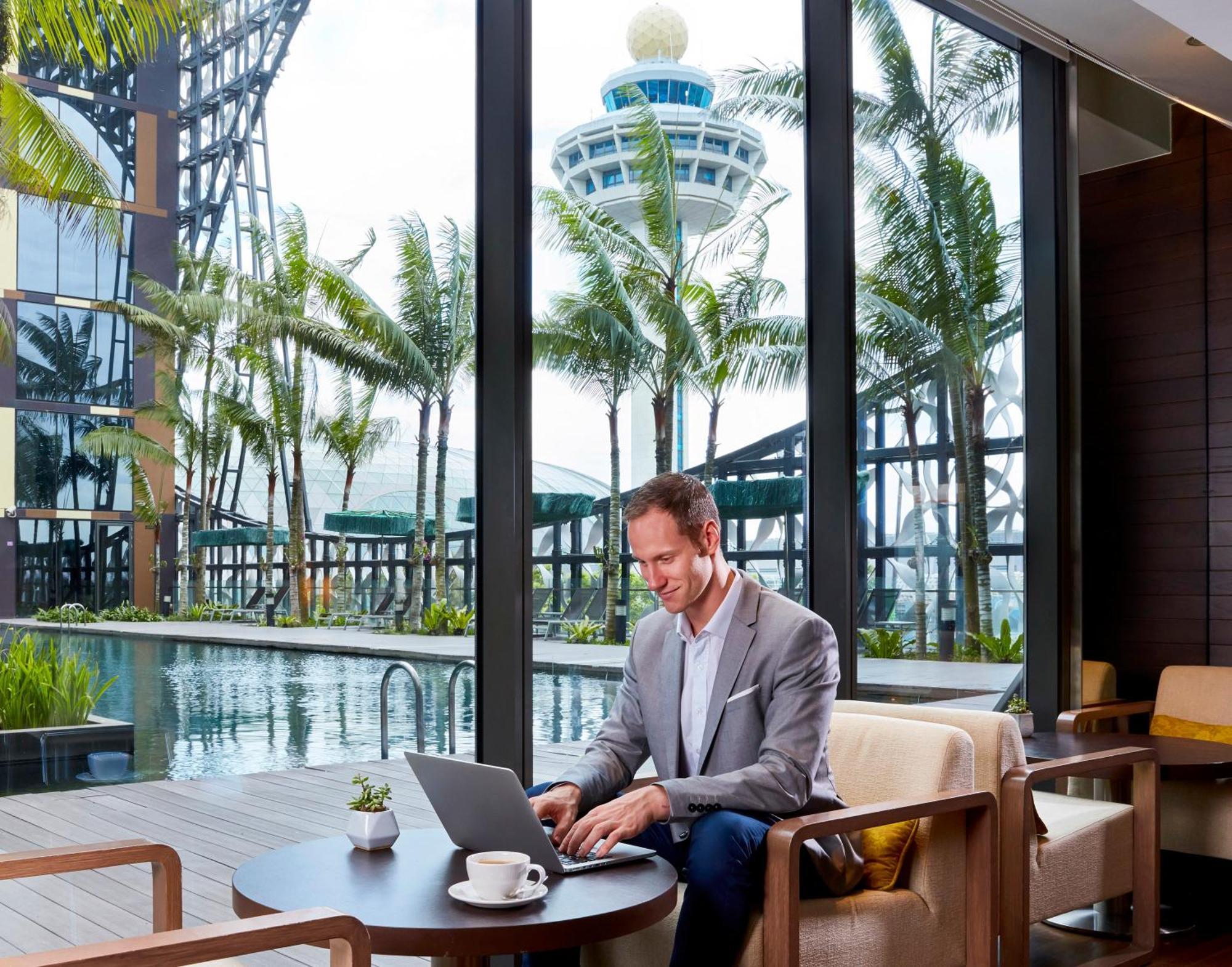 Crowne Plaza Changi Airport, An Ihg Hotel Singapore Exterior photo The photo depicts a man sitting at a modern table working on a laptop. He is dressed in a smart, light gray blazer and appears focused on his task. The setting has large windows that offer a view of palm trees and a pool, suggesting a luxurious and t