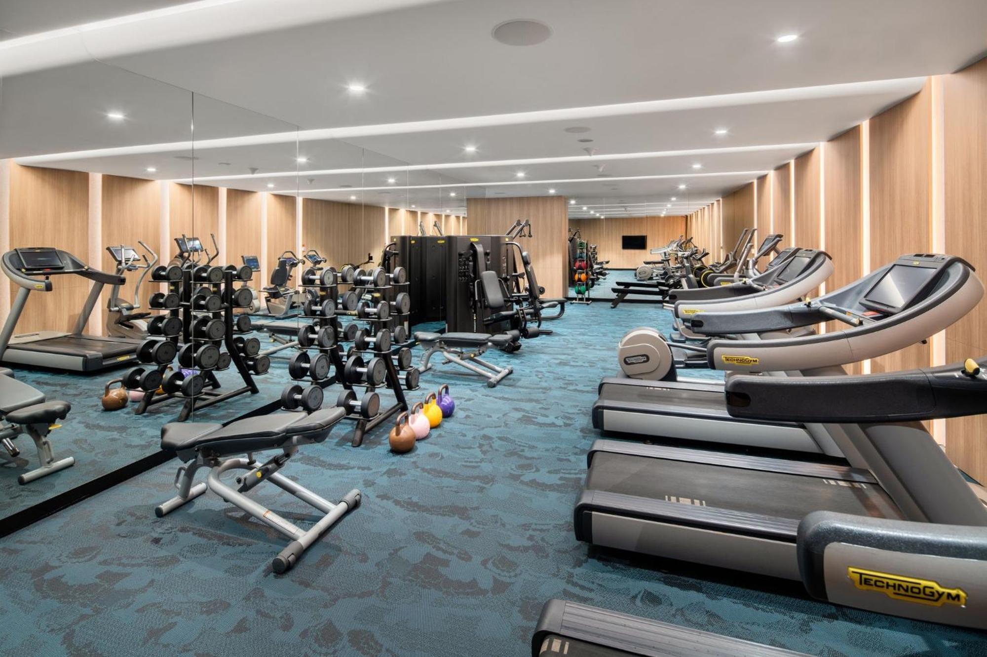 Crowne Plaza Changi Airport, An Ihg Hotel Singapore Exterior photo The photo shows a modern gym interior. It features various fitness equipment, including multiple treadmills, weightlifting machines, and a set of free weights on racks. The flooring has a blue carpet, and the walls are made of wood paneling, giving t