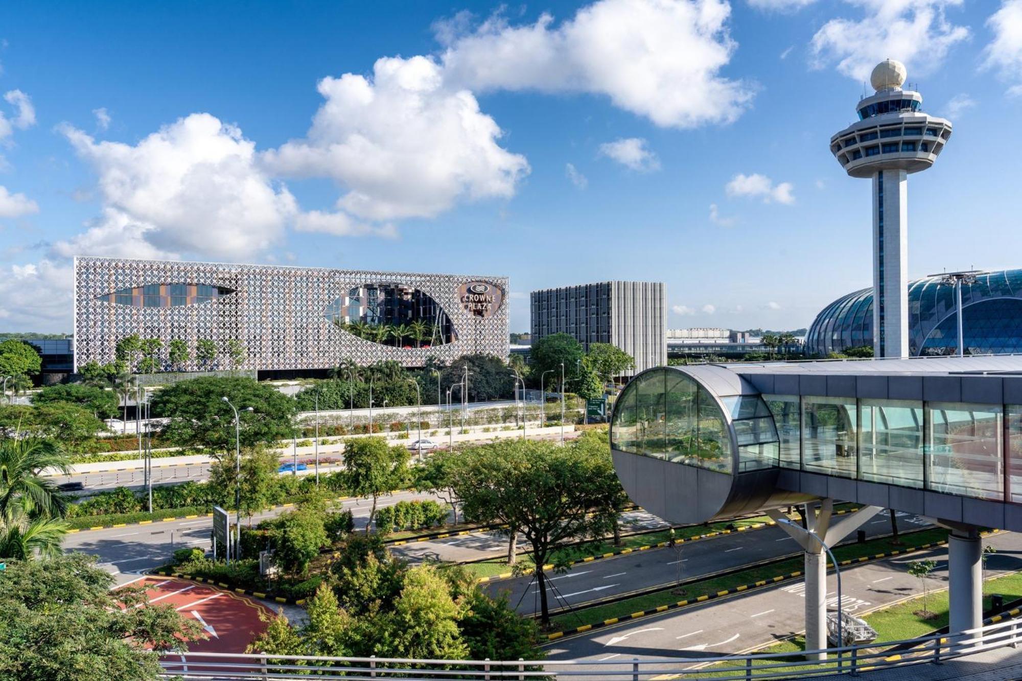 Crowne Plaza Changi Airport, An Ihg Hotel Singapore Exterior photo The photo shows a modern urban landscape with a clear blue sky and fluffy white clouds. In the foreground, there is a landscaped area with greenery and a road. Prominently featured is an interesting architectural structure, likely a building with a u