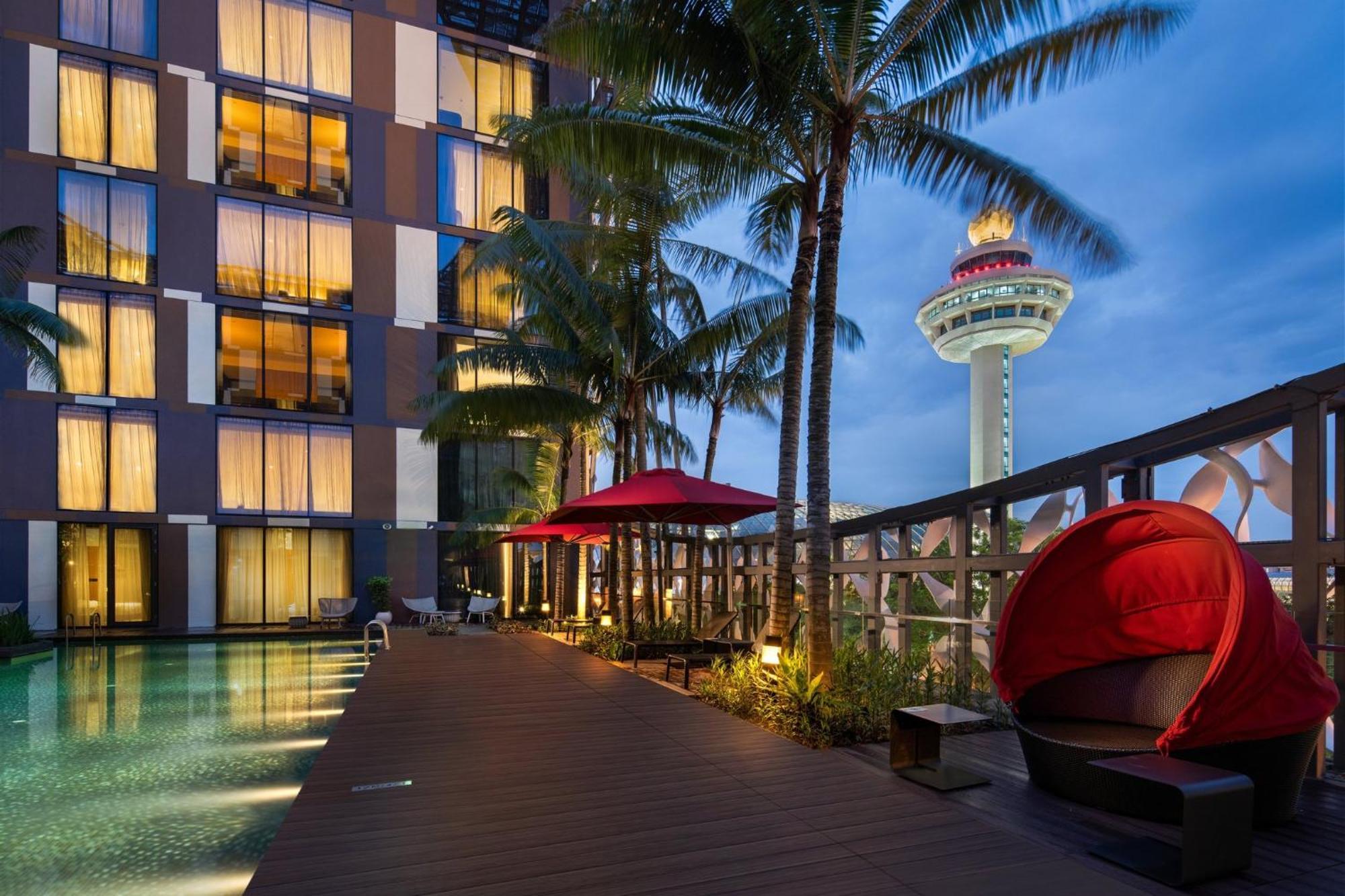 Crowne Plaza Changi Airport, An Ihg Hotel Singapore Exterior photo The image shows a modern hotel property situated near a swimming pool. The hotel features large windows with warm lighting, and it is surrounded by palm trees. There are red umbrellas providing shade, and a unique red seating area is visible near the