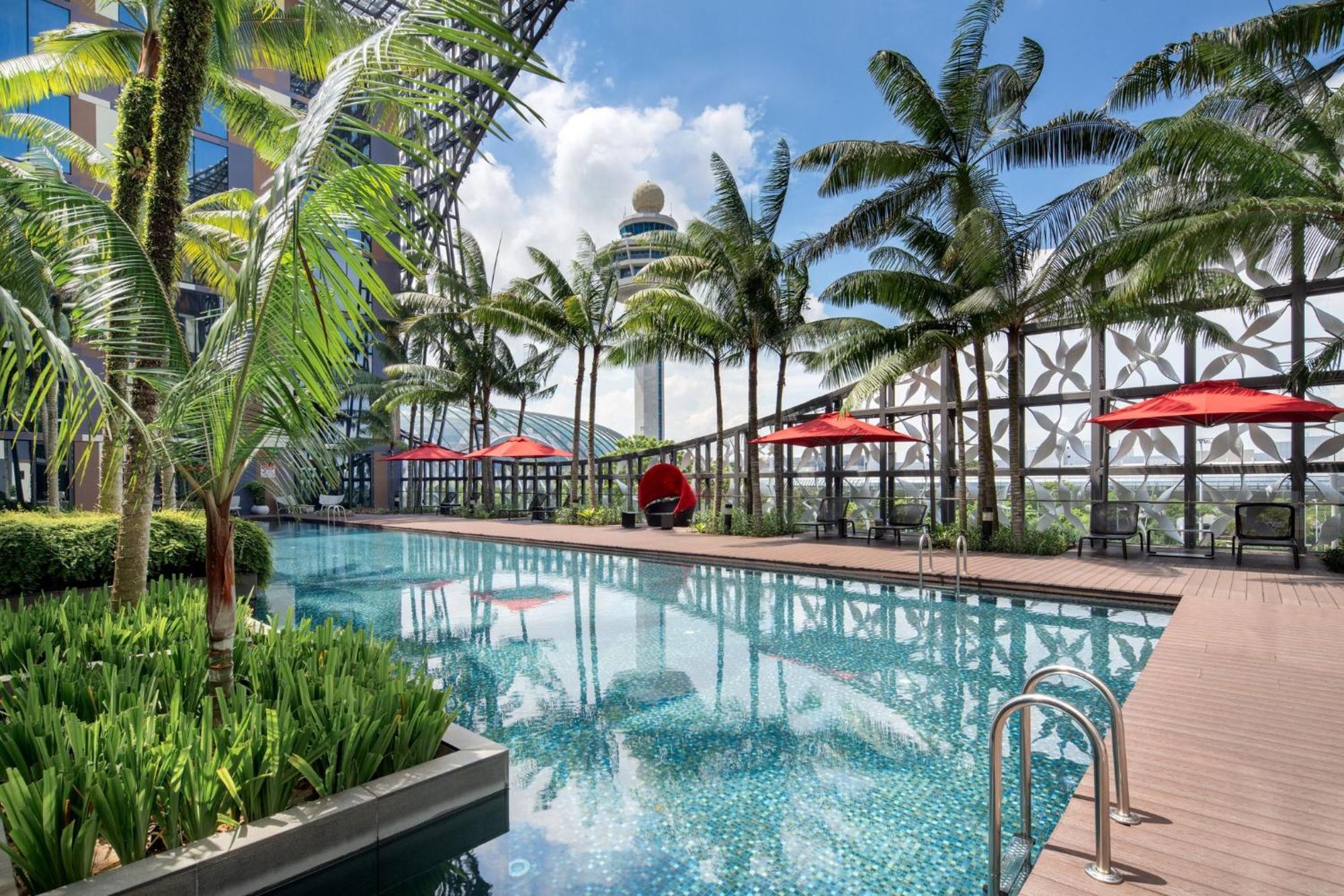 Crowne Plaza Changi Airport, An Ihg Hotel Singapore Exterior photo The image shows a luxurious pool area surrounded by palm trees. The pool has a clear blue water surface, with small tiles reflecting light. On one side, there are lounge chairs with red umbrellas providing shade. There are also some decorative plants